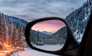 Im Seitenspiegel eines Autos sind schneebedeckte Berge und ein Sonnenuntergang zu sehen.