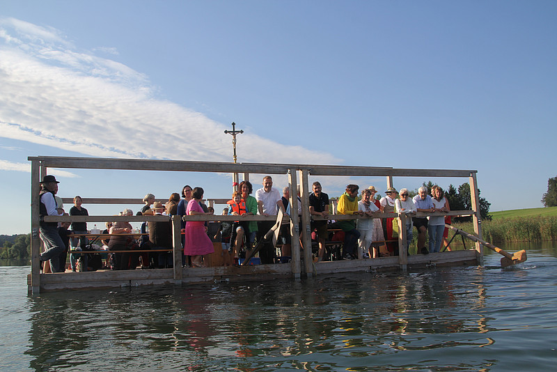 Viele Menschen verschiedenen Alters sitzen gemeinsam auf einem Floß. Mit Hintergrund ein großes Kreuz, dazu der See.