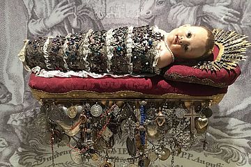 Augustinerkindl: Figur ist mit kostbaren Spitzen und Brokatbinden gefatscht, also umwickelt, auf die Perlen und bunte Glassteine genäht sind. 
