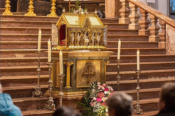 Goldener Schrein in Kirche, umstellt von brennenden Kerzen und einem Blumenkranz.
