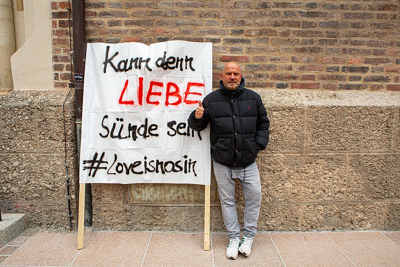 Stefan Alof steht vor dem Liebfrauendom. Neben sich ein Plakat mit der Aufschrift: "Kann denn Liebe Sünde sein?" Er erhebt seinen rechten Daumen.
