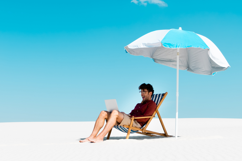 Mann mit Laptop unterm Sonnenschirm im Liegestuhl am Strand