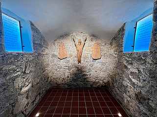 Ein kleiner Raum mit Steinwänden, an dessen Stirnseite aus Holz geschnitzte Figuren hängen. Rechts und links hängen beschriftete Tafeln an den Wänden.